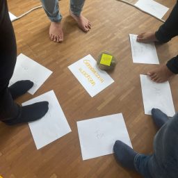 Zettel Boden Füße stehen auf Papiersheets im Kreis, in der Mitte liegt ein gelbes Würfel.